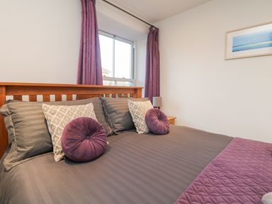 A bedroom with a wooden bed frame gray bedding purple round pillows and curtains at Miner's Rest in Troon near Camborne