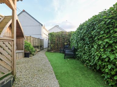 An outdoor garden area with a gravel path artificial grass black chairs wooden fence and green bushes at Miner's Rest in Troon near Camborne
