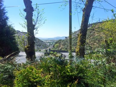 A roadside view with trees, bushes, rocky hills and a distant village at Stiniog Lodge in Blaenau Ffestiniog