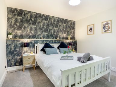 A bedroom with a white bed and blue pillows two bedside tables with lamps and plants a textured wall behind the bed and two framed pictures on a plain wall at Stiniog Lodge in Blaenau Ffestiniog