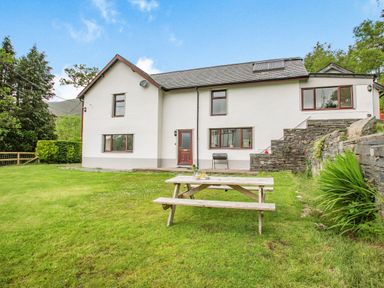 A white house with multiple windows a door and a wooden picnic table on a grassy lawn with stone steps and greenery at Stiniog Lodge in Blaenau Ffestiniog