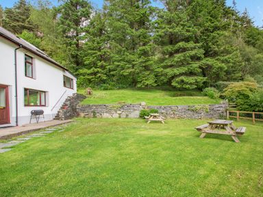 A lawn with two wooden picnic tables near a white house with stone steps and trees in the background at Stiniog Lodge in Blaenau Ffestiniog