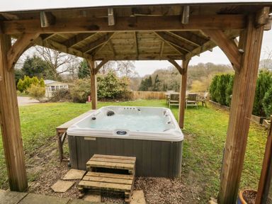 An outdoor hot tub under a wooden gazebo with wooden steps and garden furniture on a grassy lawn at Shatterford Lakes in Shatterford