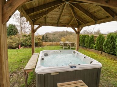 A covered outdoor area with a hot tub and wooden bench in a garden with wooden table and chairs at Shatterford Lakes in Shatterford