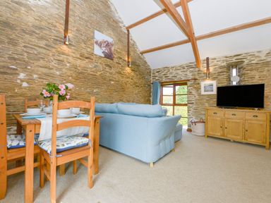 A living room with a wooden dining table and chairs a blue sofa a wooden cabinet with a television and stone walls at Erin Cottage in Pelynt near Looe