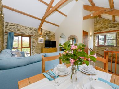 A dining table with flowers and place settings overlooking a living room with a blue sofa and exposed stone walls at Erin Cottage in Pelynt near Looe