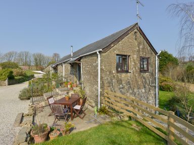 An outdoor seating area with a wooden table and chairs beside a stone cottage with potted plants and a gravel driveway at Erin Cottage in Pelynt near Looe