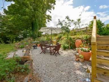 An outdoor garden seating area with a table and chairs under a large umbrella surrounded by plants and gravel at Erin Cottage in Pelynt near Looe