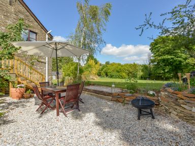 An outdoor patio with a wooden dining table and chairs under a white umbrella and a fire pit near a stone wall at Erin Cottage in Pelynt near Looe
