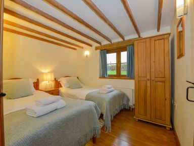 A bedroom with two single beds with folded towels on them a bedside table with a lamp a wooden wardrobe and a window with blue curtains at Hook Cottage in Pelynt near Looe