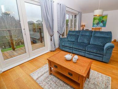 A living room with a blue sofa a wooden coffee table with a tea set and glass doors with curtains at 1 Lake View in Lanreath