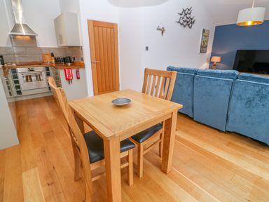 A dining area with a wooden table and two chairs next to a kitchen and a living room with blue sofas at 1 Lake View Lanreath
