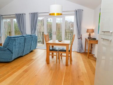 A dining area with a wooden table and chairs next to a blue sofa and large windows with gray curtains at 1 Lake View in Lanreath