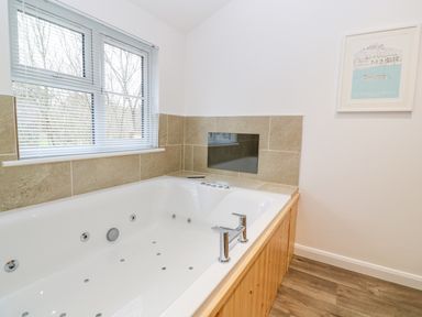 A bathroom with a large white jacuzzi tub wood paneling around the tub tiled walls a window with blinds and a framed picture on the wall at 1 Lake View in Lanreath