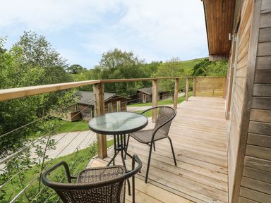 A wooden balcony with a glass table and two chairs overlooking trees and wooden cabins at 1 Lake View in Lanreath