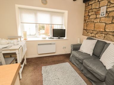 A small living room with a gray sofa and cushions a dining table with tea set a window with a TV on the sill and a stone wall at Wren Cottage in Morpeth