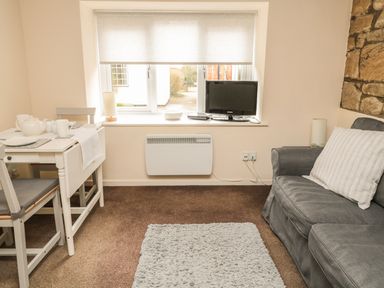 A small living room with a gray couch a white dining table with chairs a window with blinds and a television on the windowsill at Wren Cottage in Morpeth