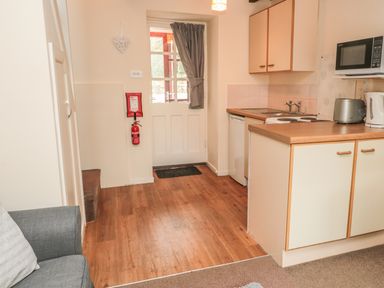 A small kitchen area with wooden flooring a white door with a curtain a gray sofa and kitchen cabinets with microwave toaster and kettle at Wren Cottage Morpeth
