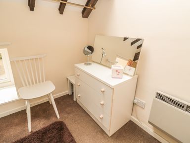 A white chair next to a white dresser with a mirror and a small round mirror on top in a room with beige walls at Wren Cottage in Morpeth