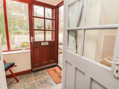 An entryway with a partially open white door showing a red wooden door with glass panels and a welcome mat at Wren Cottage in Morpeth