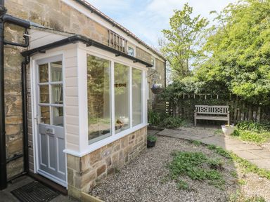 A stone house with a white door and windows next to a garden with a wooden bench and plants at Wren Cottage in Morpeth
