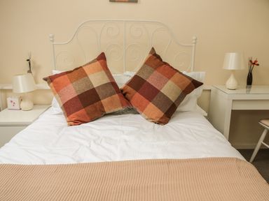 A bed with plaid cushions and white bedding with bedside tables and lamps at Heron Cottage in Morpeth