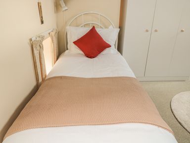 A bedroom with a single bed with white sheets a beige blanket and a red pillow next to a white wardrobe in Heron Cottage Morpeth