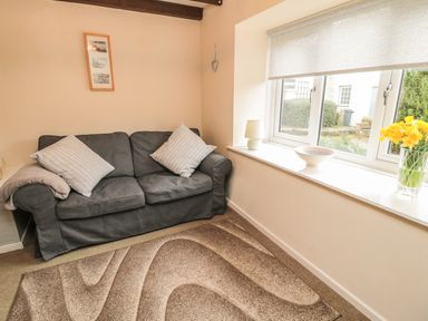 A small living room with a gray sofa two pillows a patterned rug and a windowsill with a lamp bowl and vase of yellow flowers at Heron Cottage in Morpeth