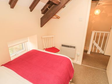 A small bedroom with a bed covered by a red blanket a white chair with a red cushion and a window under wooden ceiling beams at Magpie Cottage in Morpeth