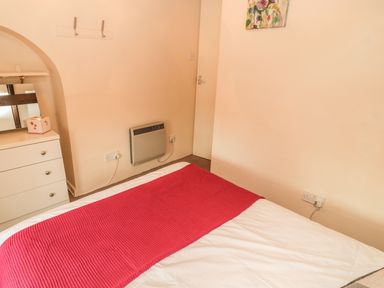 A bedroom with a bed covered with a red throw a white dresser with a mirror a wall-mounted heater and a floral painting at Magpie Cottage in Morpeth