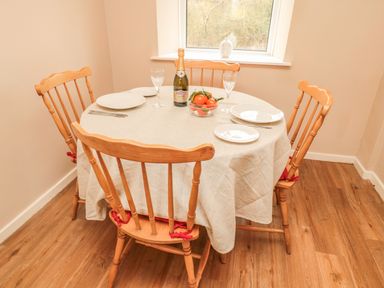 A dining table with four wooden chairs set with plates glasses a bowl of tomatoes and a bottle under a window at Magpie Cottage in Morpeth
