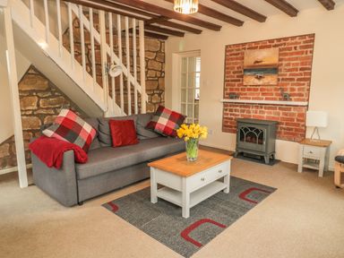 A living room with a gray sofa with red and checkered cushions a wooden coffee table with yellow flowers a brick fireplace and a white door at Magpie Cottage in Morpeth