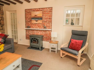 A living room with a black armchair and red cushion a grey stove with a painting above it and a wooden coffee table at Magpie Cottage in Morpeth