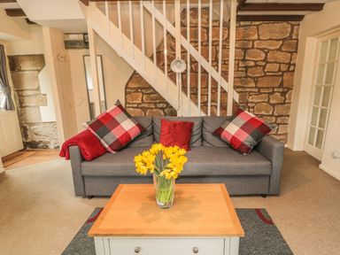 A living room with a gray sofa and red checkered cushions a wooden coffee table with yellow flowers and stone wall at Magpie Cottage in Morpeth