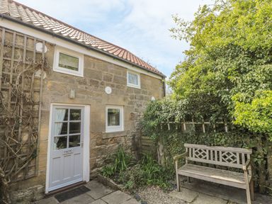 Stone cottage exterior with white door and windows garden bench and greenery at Magpie Cottage in Morpeth