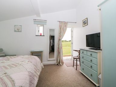 A bedroom with a bed covered with a quilt a dresser with a television a chair a small table a wall mirror and a door leading outside at Owl Barn in Tavistock