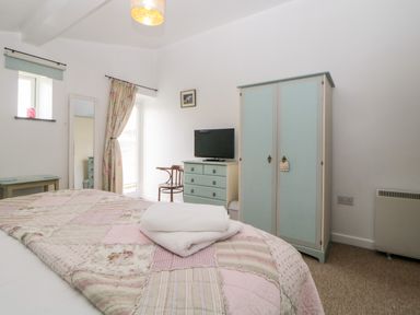 A bedroom with a bed covered with a quilt and towels a blue wardrobe blue chest of drawers with television a chair a tall mirror and a window with curtains at Owl Barn in Tavistock