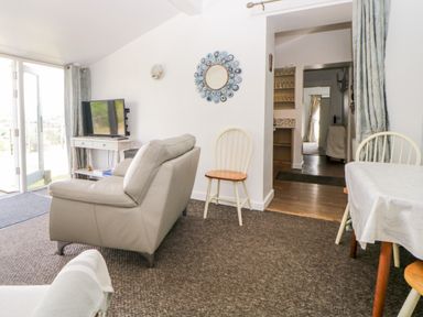 A living room with a TV on a white table a grey sofa wooden chairs and a dining table at Owl Barn in Tavistock