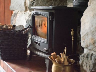A wood burning stove with firewood in a basket next to a stone wall at Cae Lleci in Llanfachreth near Dolgellau
