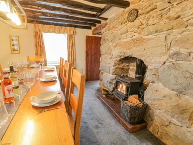 A dining room with a wooden table and chairs a stone wall with a wood burning stove and a basket of firewood at Cae Lleci in Llanfachreth near Dolgellau