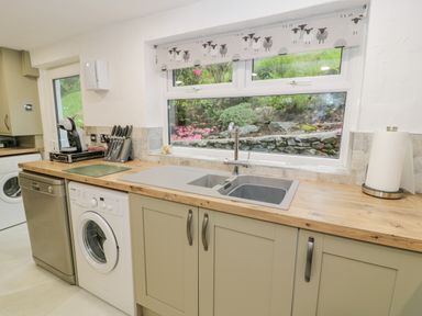 A kitchen countertop with a sink, dishwasher, washing machine, knife set, coffee machine and window with a floral view at Cae Lleci in Llanfachreth near Dolgellau
