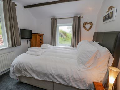 A bedroom with a double bed with white bedding a wooden dresser a wall-mounted television and two windows with curtains at Cae Lleci in Llanfachreth near Dolgellau