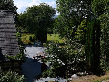 An outdoor patio area with a round wooden picnic table and benches near a house roof with slate tiles surrounded by trees and greenery at Cae Lleci in Llanfachreth near Dolgellau