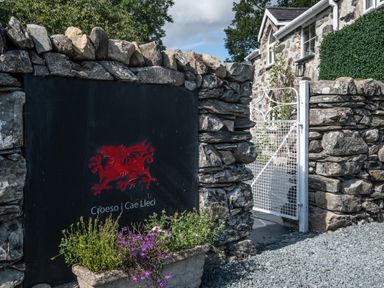 A stone wall with a black sign featuring a red dragon and Welsh text beside a white metal gate at Cae Lleci in Llanfachreth near Dolgellau