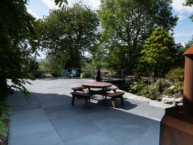 An outdoor patio with a round wooden picnic table in the center two blue chairs and a small dining table with three chairs in the background surrounded by trees and plants at Cae Lleci Llanfachreth near Dolgellau