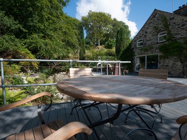 An outdoor wooden table with chairs on a terrace next to a stone house surrounded by trees at Cae Lleci in Llanfachreth near Dolgellau