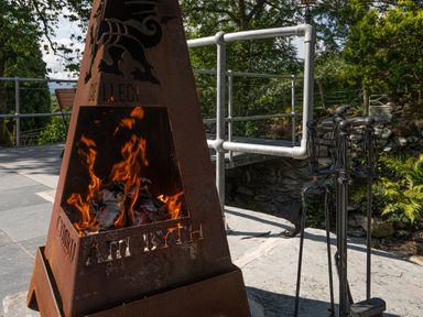 An outdoor metal fire pit with flames and fire tools next to it on a stone patio at Cae Lleci Llanfachreth near Dolgellau