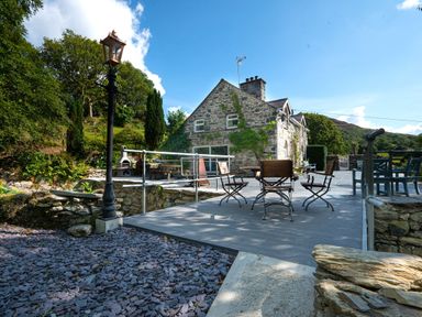 An outdoor patio with wooden chairs and tables next to a stone house surrounded by greenery at Cae Lleci in Llanfachreth near Dolgellau