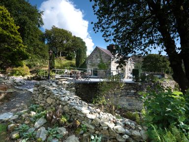 A stone house with a raised patio and outdoor furniture surrounded by stone walls and trees at Cae Lleci Llanfachreth near Dolgellau