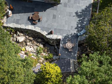 An outdoor stone patio with round wooden tables and chairs surrounded by trees and garden at Cae Lleci Llanfachreth near Dolgellau